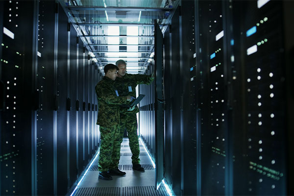 Two soldiers standing in a sever room, one of them holding a laptop.