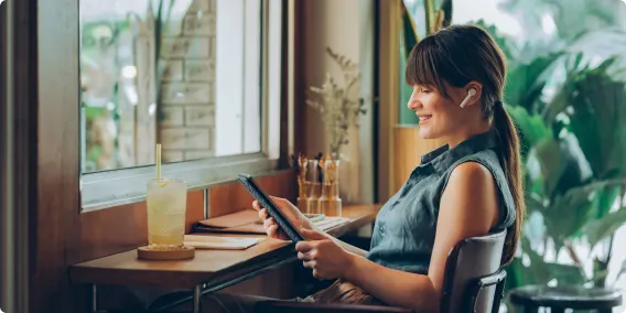 A smiling woman sitting at a desk, wearing earbuds and looking at a tablet.