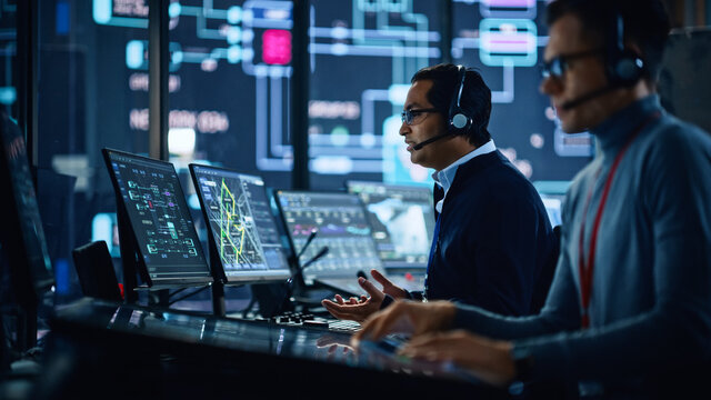 Portrait of Professional IT Technical Support Specialist Working on Computer in Monitoring Control Room with Digital Screens. Employee Wears Headphones with Mic and Talking on a Call.