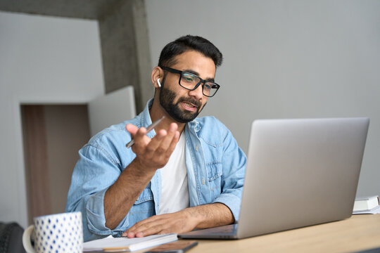 Young eastern indian Hispanic male teacher wearing glasses with earphones having webinar video training class using laptop at modern home office. Remote distant online work e learning tech concept.