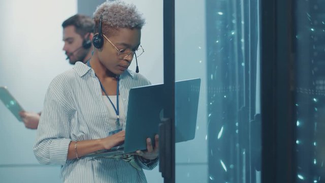 African woman IT support worker using a laptop for server diagnostics, inspecting supercomputer hardware, working with male specialist in high tech datacenter.