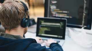 Male software developer using AI coding tools and writing code on a laptop while wearing headphones in an office space.