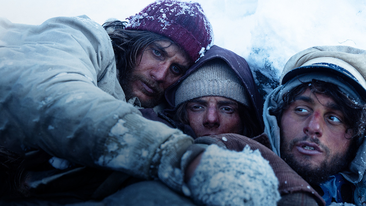 Three men gather closely laying in the snow with concerned expressions in 'Society of the Snow.'