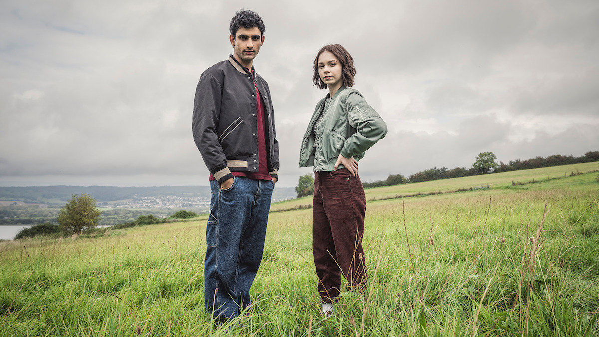 Zain Iqbal as Ravi Singh and Emma Myers as Pip Fitz-Amobi stand on a grassy hill together in ‘A Good Girl’s Guide to Murder.’