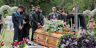 Sebastian Dante as Alex, Eréndira Ibarra as Lupita, and Alberto Guerra as El Charro stand in front of a casket surrounded in floral arrangements at a cemetery.