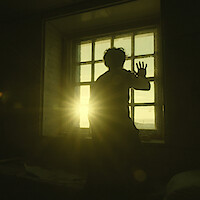 A man puts his hand to a window as the sun rises in the distance.