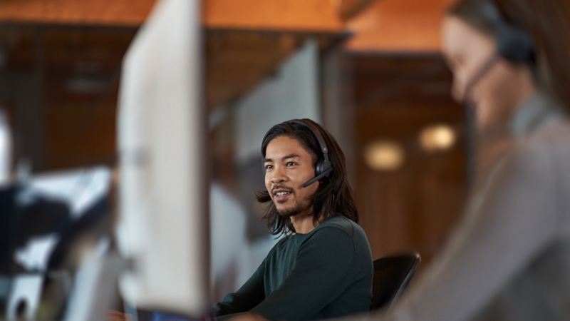 Mand med langt hår med headset, der arbejder på en computer på et kontor med en kollega i baggrunden