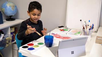 A young boy uses paints on paper as he watches an open Surface laptop