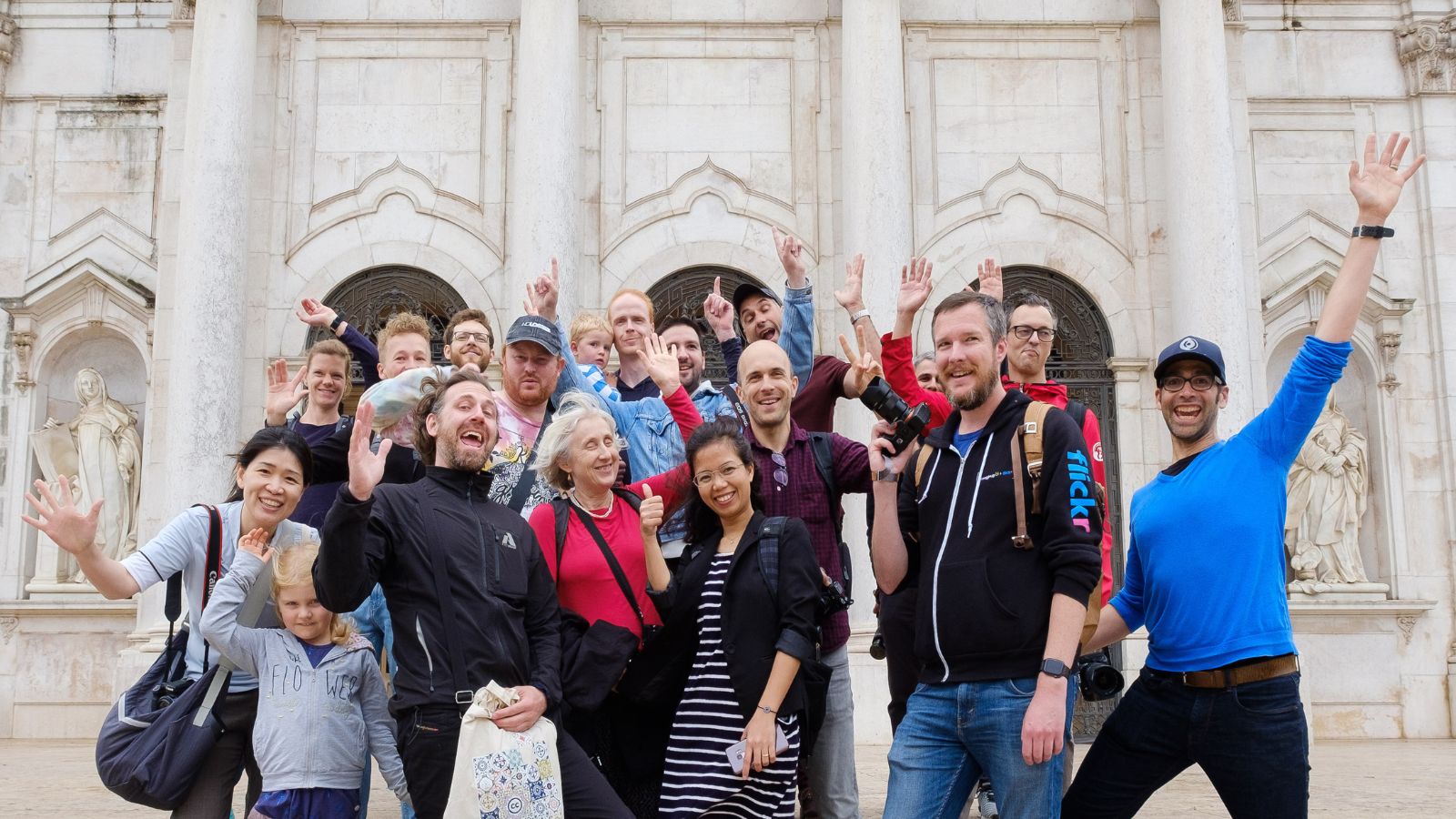 A group of about 18 people of all ages smiling and waving in front of the doors of a white historical building.