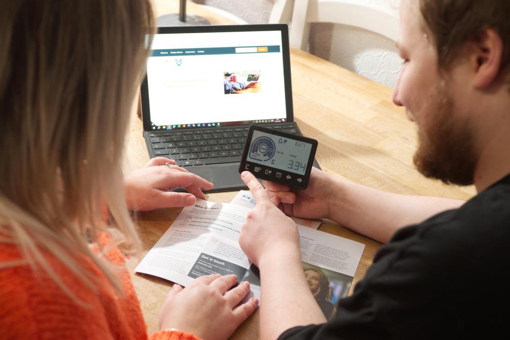 A customer sits infront of her computer with a leaflet on the table and an advisor next to her holding a smart meter and showing her how it works