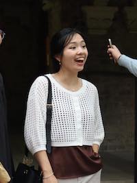 Marian standing amd smiling while wearing a white cardigan and brown camisole.