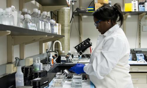 A woman in a lab, wearing a white lab coat and gloves. 