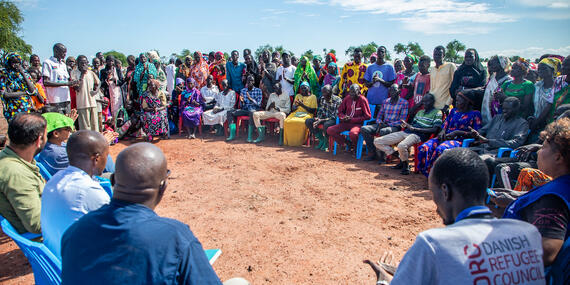 People sit in chairs facing each other, some people can also been standing behind them.