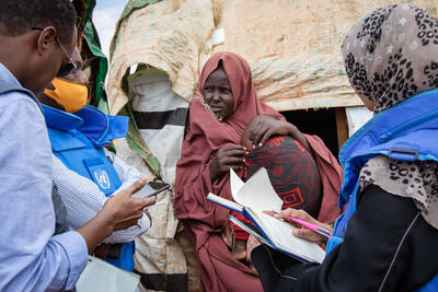 A woman with baby in her arms stands in front of an informal tent. Two men and a woman in blue vests and clothing face her.