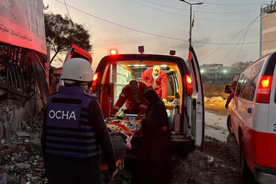 A man wearing an OCHA vest and helmet is helping upload an ambulance stretcher with a person lying on it into an ambulance, which has its back doors open. Another ambulance can be seen parked alongside it.