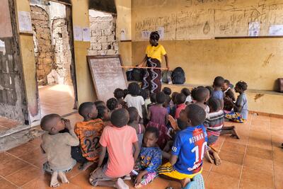 A woman stands with a pointer and watches as a child writes on a blackboard placed against a wall. Children are seated on the floor in front of the board. Soe bags can be seen placed against a dilapidated wall.