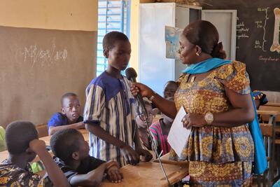 Inside a classroom, a boy standing behind a desk speaks into a microphone, which held by a woman, who is standing in front of him. Other children can be seen seated at their desks. Two blackboards are in view in the background.