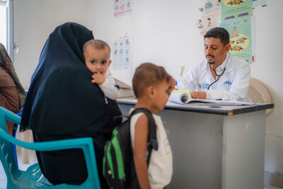 A man in a white jacket with a stethoscope sits behind a desk - a woman with a baby in her arms and a young boy standing next to her sits across the desk.