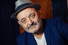 Homme ridé aux cheveux gris et barbe, de face avec un chapeau.