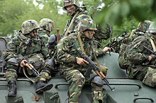 Six soldiers dressed in camouflage and face paint sit on top of a green vehicle.