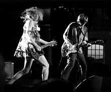 A woman and a man playing guitar in performance. The woman on the left is dressed in a short dress and the man on the right is in jeans and a shirt.