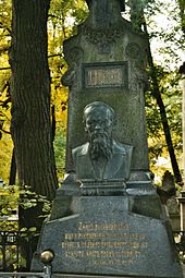 Une pierre tombale dans un chemin boisé. Dessus, un buste d'homme chatoyant.