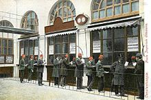 Ten men in suits, most in red fezes, wait before three ticket windows. Over them, two clockfaces. The postcard has the French text "Constantinople. Poste Impériale Ottomane.
