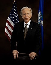 A man in a formal suit standing in front of national and state flags.