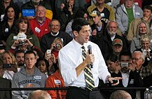 A middle-aged man in a white shirt and black and yellow striped tie speaks into a microphone on stage in front of a crowd.