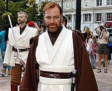 Homme barbu portant un vêtement blanc et un manteau marron.