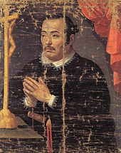 Painting of an Asian man in black priest robes with white collar praying in front of a crucifix.