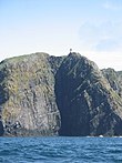 A small white building is barely visible on top of dark and imposing cliffs with deep blue water at their base.