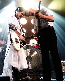 Michelle Zauner and Peter Bradley standing and playing guitar while resting the tops of their heads against each other