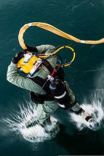 A US Navy surface supplied diver wearing a lightweight demand helmet and holding the umbilical at head level is shown entering the water by jumping in. The view is from the deck from which the diver has jumped, and shows the back of the diver as the fins first contact the water