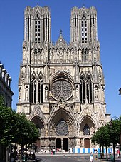 Frontale Farbfotografie einer gotischen Kirche mit zwei breiten, rechteckigen Turmspitzen. Viele kleine Türme, Säulen und Figuren verzieren das gesamte Gebäude. In der Mitte ist eine große Rosette, die von vier langen, schmalen Säulenfenstern begrenzt ist. Der Eingang besteht aus drei Spitzbögen, die mit Rosetten und vielen Figuren verziert sind.