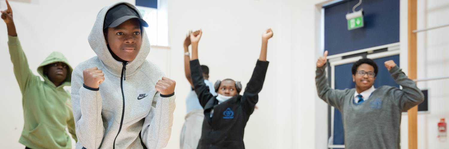 A group of young people are moving. There are four clearly visable in the image, but you get the feeling that there are more people doing the same. They have their arms raised above their heads.