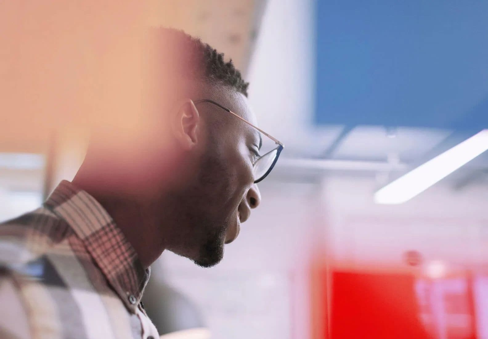 A man wearing glasses in a work environment looking downwards and smiling.