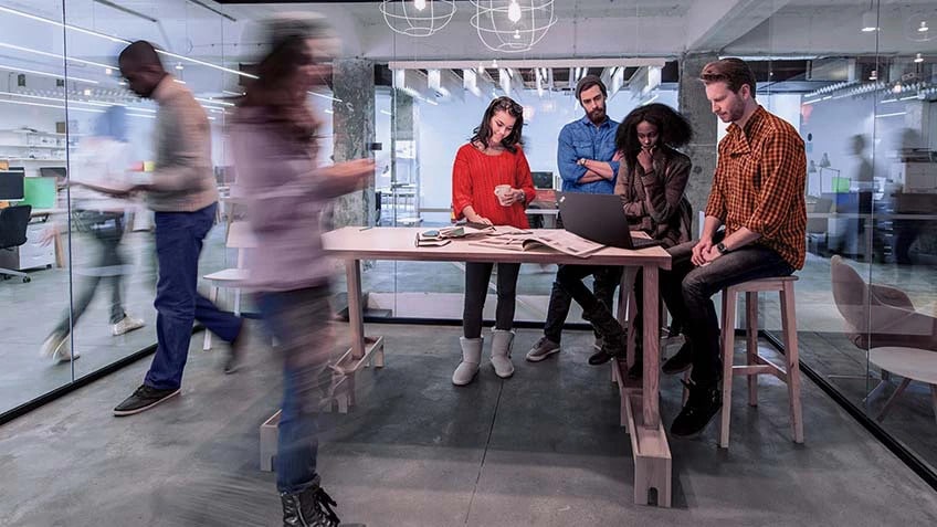 Four people meeting in an all-glass conference room with colleagues moving about around them.