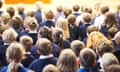 Children sat in a school hall wearing navy blue jumpers, photographed from behind