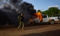 An armed man in fatigues and combat gear watches a pick-up truck burn