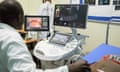 A man in a white coat passes a probe over a woman's abdomen and looks at a screen.