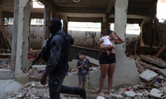 A police in a balaclava holding a large gun walks by a woman and two children in front of a destroyed building.