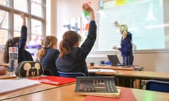 Backs of children sitting in classroom with hands up as teachers stands at white board