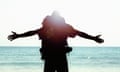 A man wearing a backpack looking out to sea with his arms outstretched.