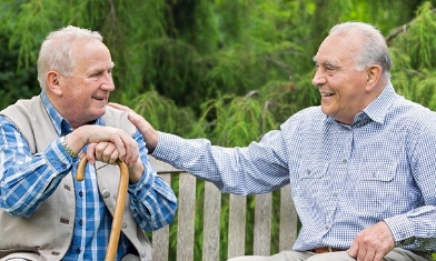 Two elderly men sat on a park bench, one with his hand on others shoulder, smiling at each other