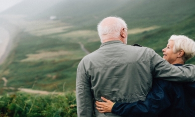 Elderly couple looking at each other lovingly, stood on a high hillside overlooking coastline below