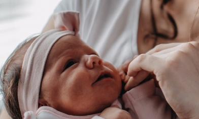 Baby girl in a pink outfit and matching headband, looking at her mum, holding her mum's hand