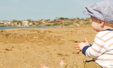 Clothed male baby wearing a flat cap sat on a beach with sand in his hands