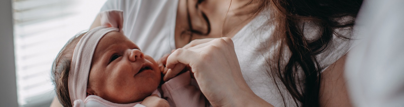 Baby girl in a pink outfit and matching headband, looking at her mum, holding her mum's hand