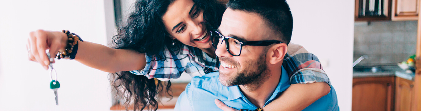 Young couple in kitchen both smiling while woman holds dangling key out in front of man